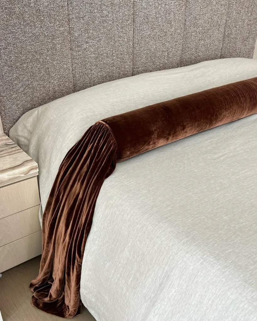 Brown velvet bolster cushion on light textured bedding against a grey upholstered headboard
