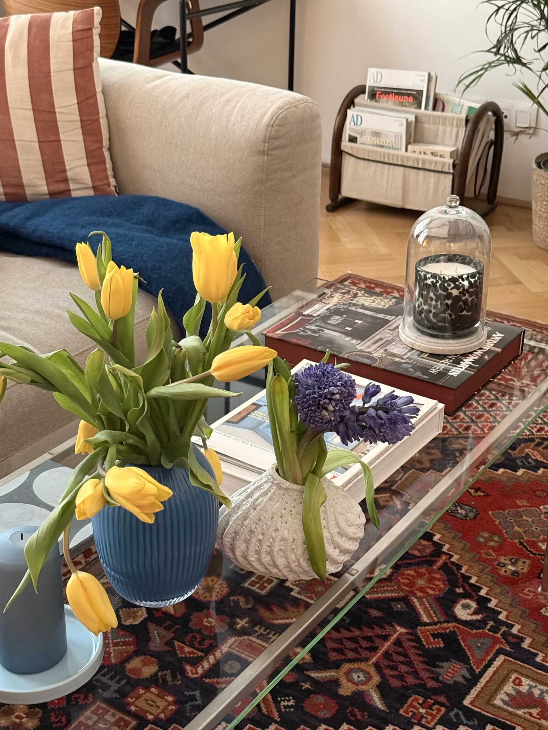 Yellow tulips in a blue vase on a glass coffee table with books and a glass cloche, Persian rug below