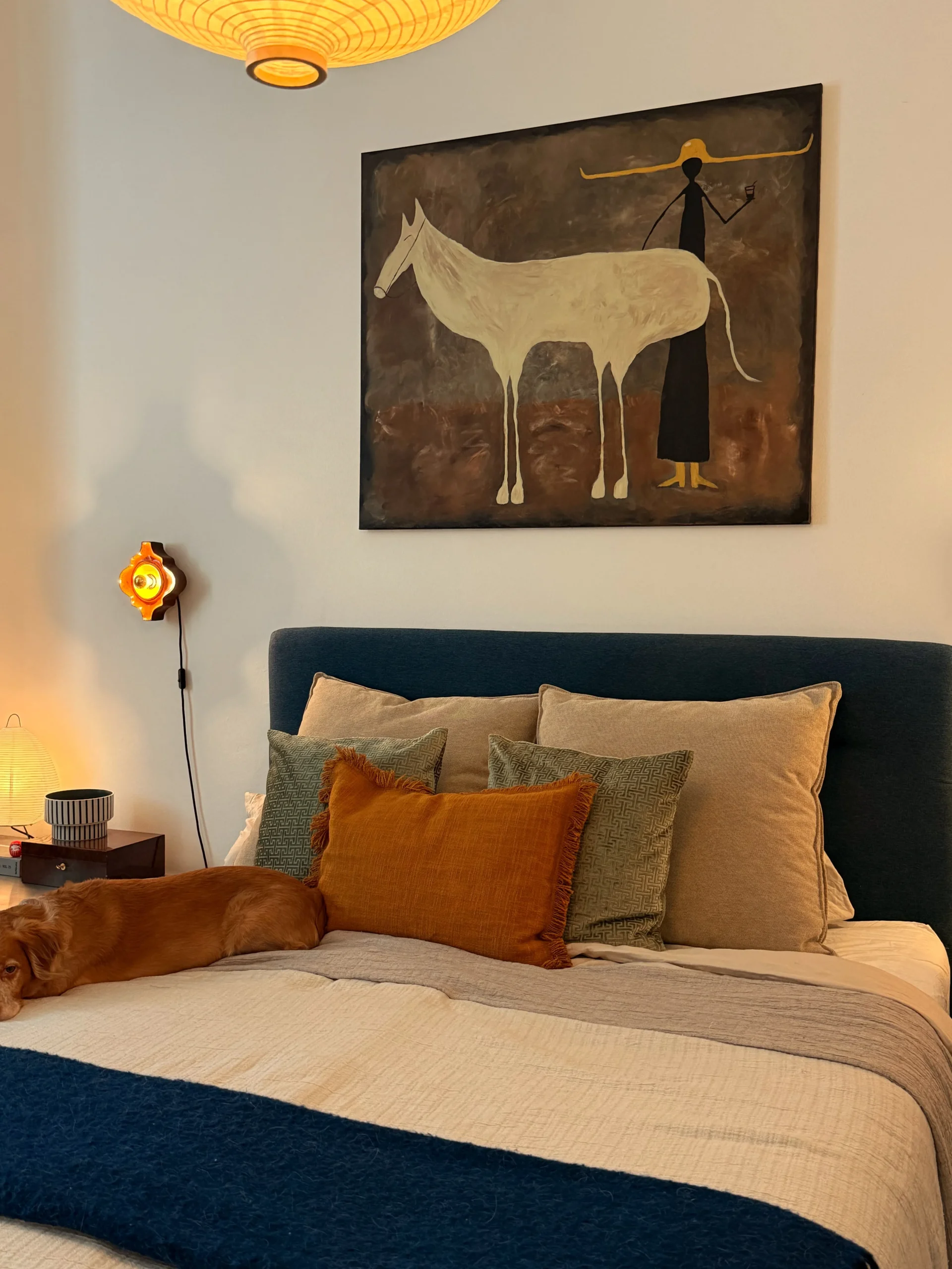 Bedroom with a navy upholstered bed, rust and sage cushions, a horse painting above and a dog on the bed