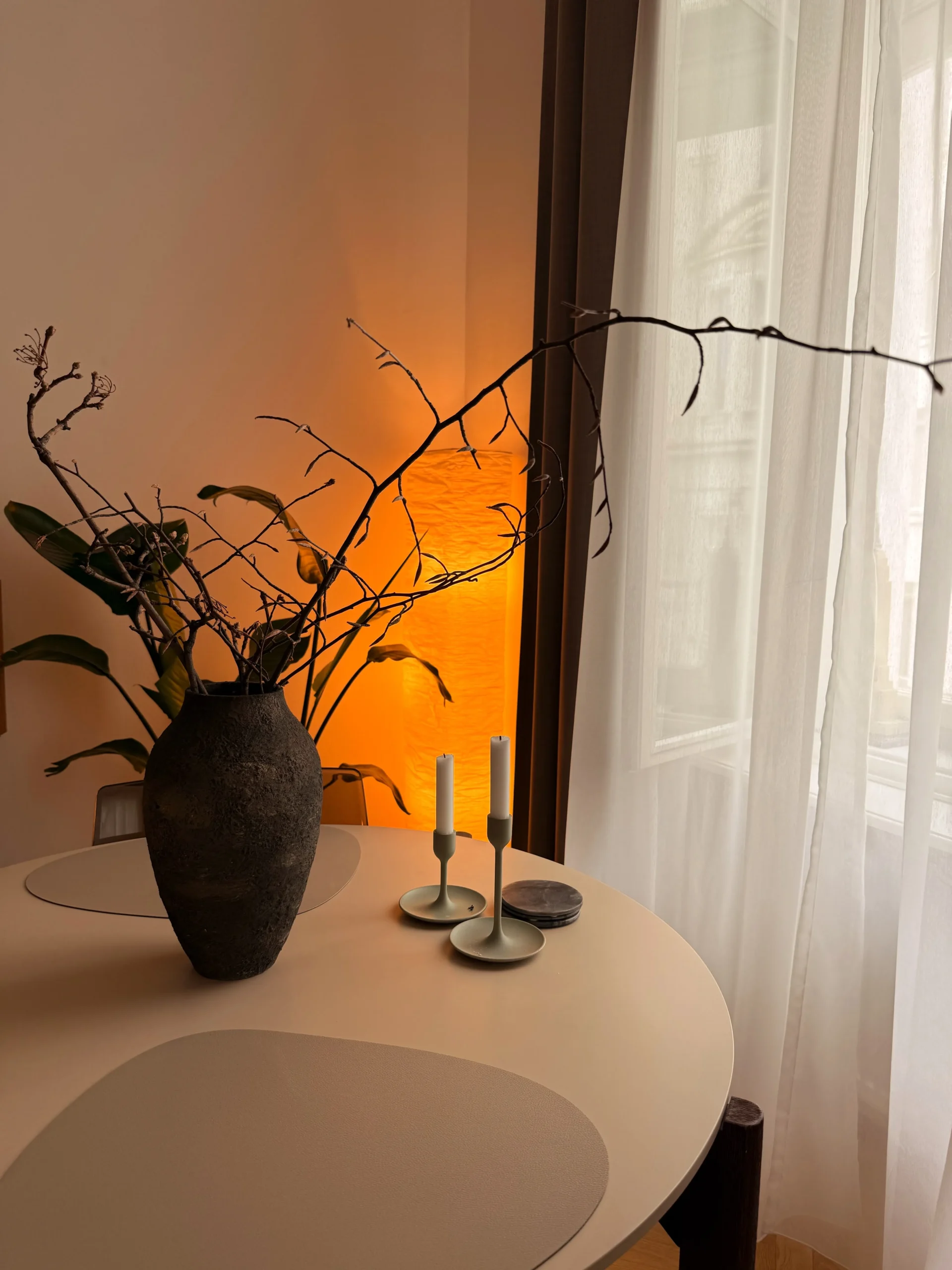 Round dining table with a dark stone vase of branches, brass candlesticks and warm lamplight by sheer curtains