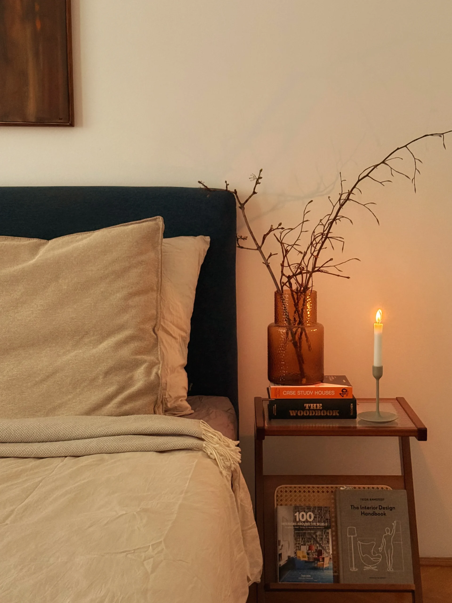 Bedside with linen bedding, a navy headboard, branches in an amber vase, books and a lit candle on a wooden side table